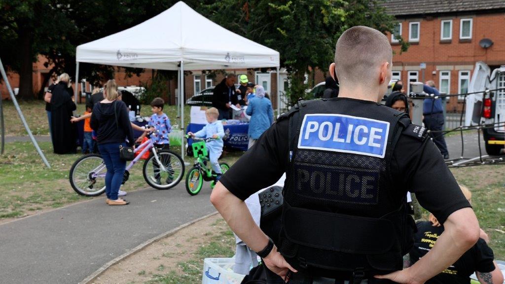 An officer stands with his back to the camera, which reads 'police', as he overlooks a communtiy engagement event in a park.