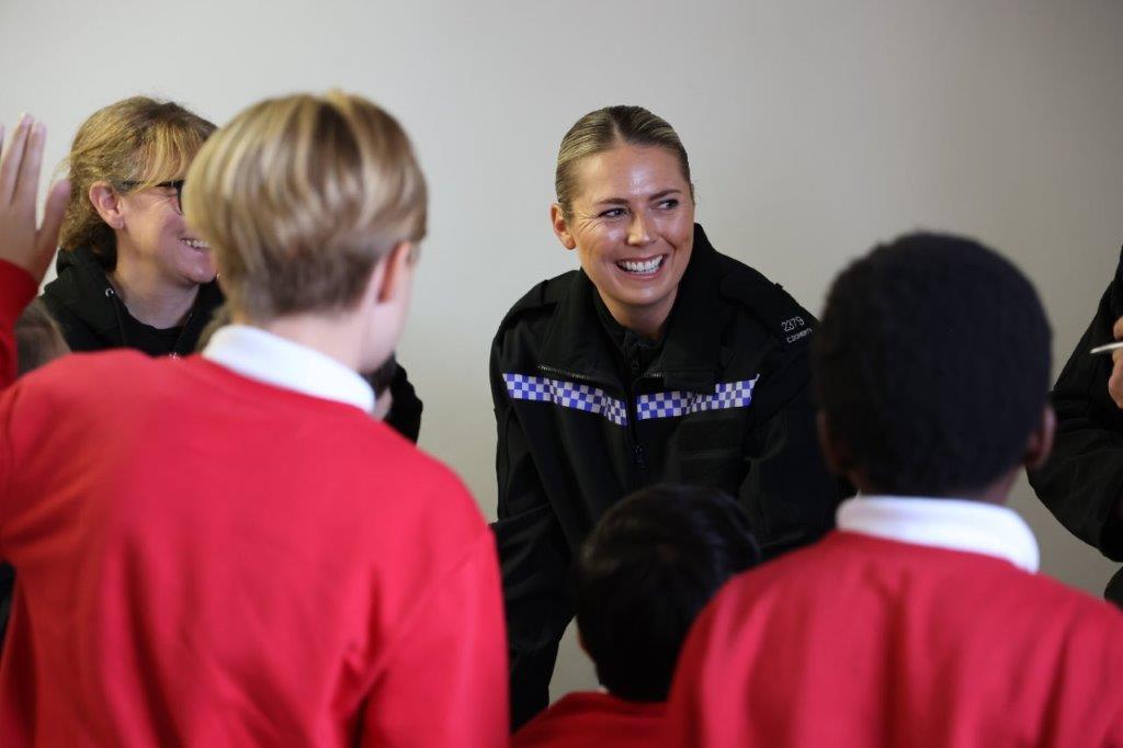 A smiling SEIO stands behind a table as she delivers an activity to a group of schoolchildren, with two pupils in the foreground.