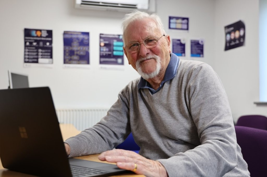 Volunteer John Duke sits at a desk with a laptop in front of him, while smiling towards the camera.