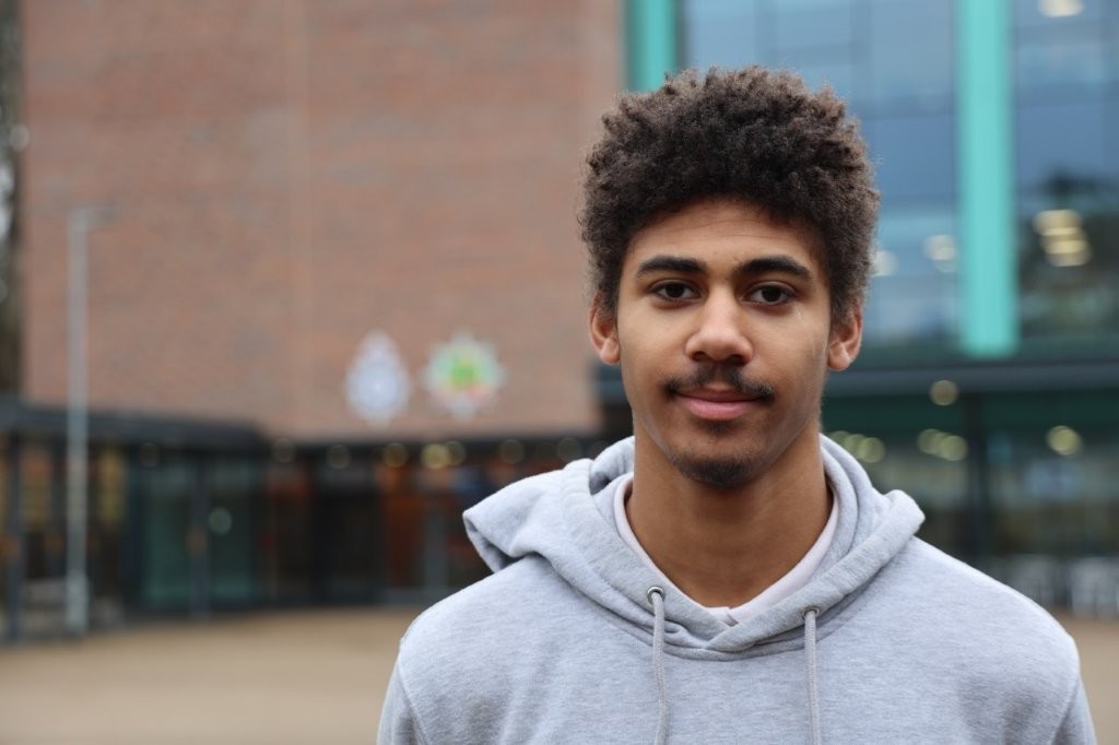 A headshot of previous Nxt Gen Award winner R'amarnai Anderson. He is stood at police HQ in front of a glass building. Wearing a grey hooded top, he has a moustache and goatee.
