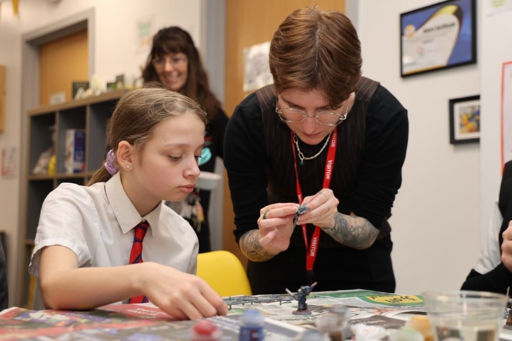 A staff member stands over the shoulder of a pupil, who is sitting at a table, to help her paint.