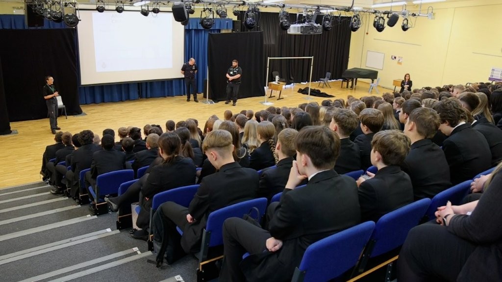 An assembly of schoolchildren listen to a presentation from a police officer.