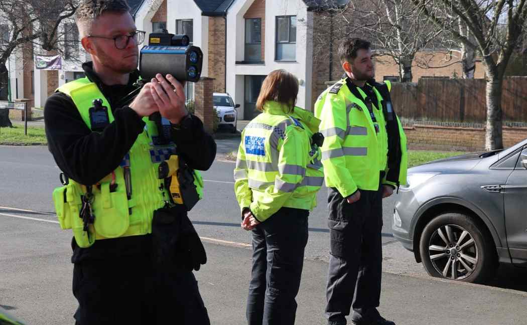 Officer stands pointing a speed gun, with two officers standing behind
