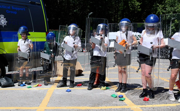 Schoolchildren stand in a line next to police van wielding riot shield and helmets as brightly-coloured bean bags are thrown at them.