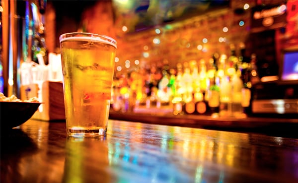A beer glass in the foreground on a bar, with the background out of focus with bottles of drink behind the bar.