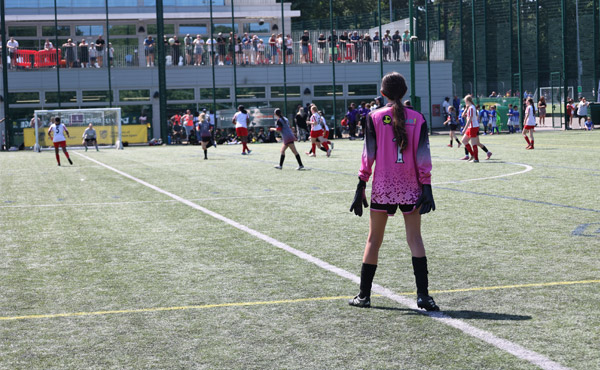 A goalkeeper stands on the edge of her box, watching play at the other end of the pitch.