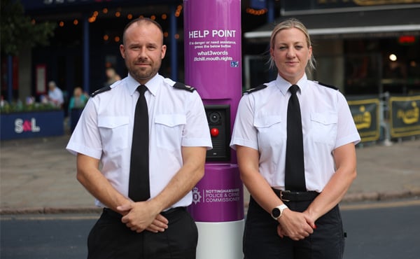 Two police officers in uniform stand either side of a new help point, a metal pole, which has a red button for people to press for help.