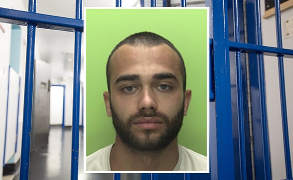 A custody photo of Tommy-John Mustafa. A man, standing in front of a lime green background. He has olive skin and is wearing a white t-shirt. He has dark brown, short hair in a buzzcut-style. He has a dark brown, full beard and goatee,  