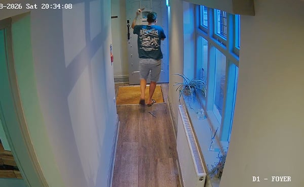 A man in shorts and a t-shire walking along a corridor with his hands in the air, as a knife falls from his back pocket.
