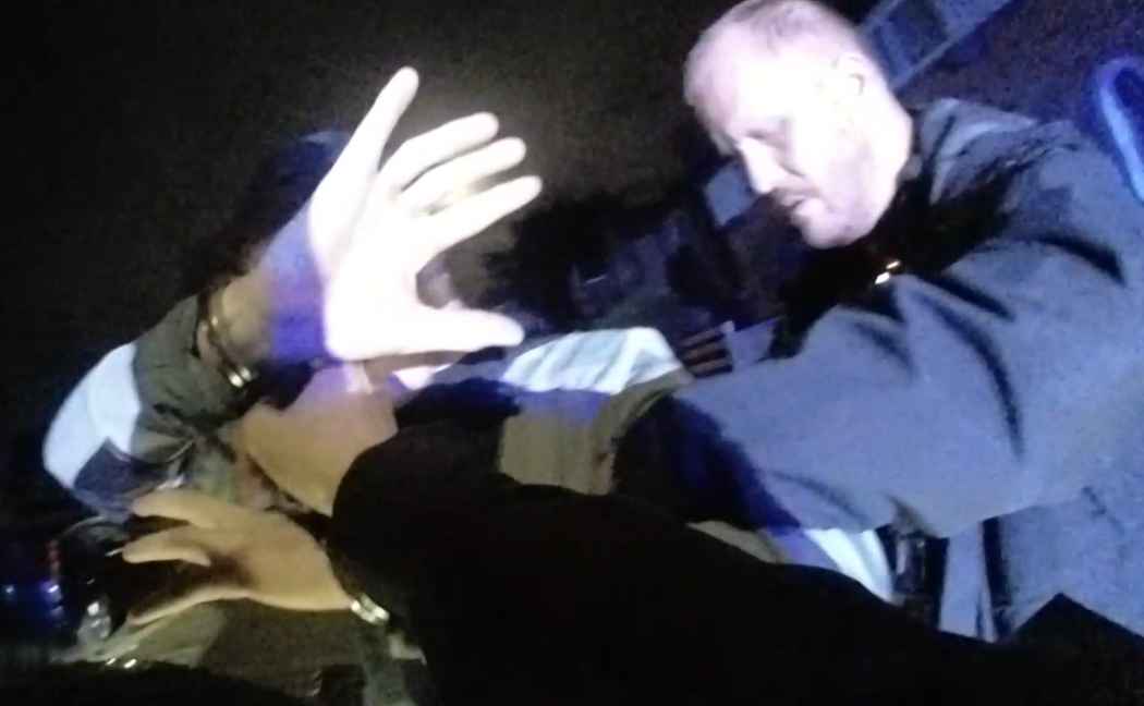 A suspect holds their hands up to the camera while being placed in handcuffs